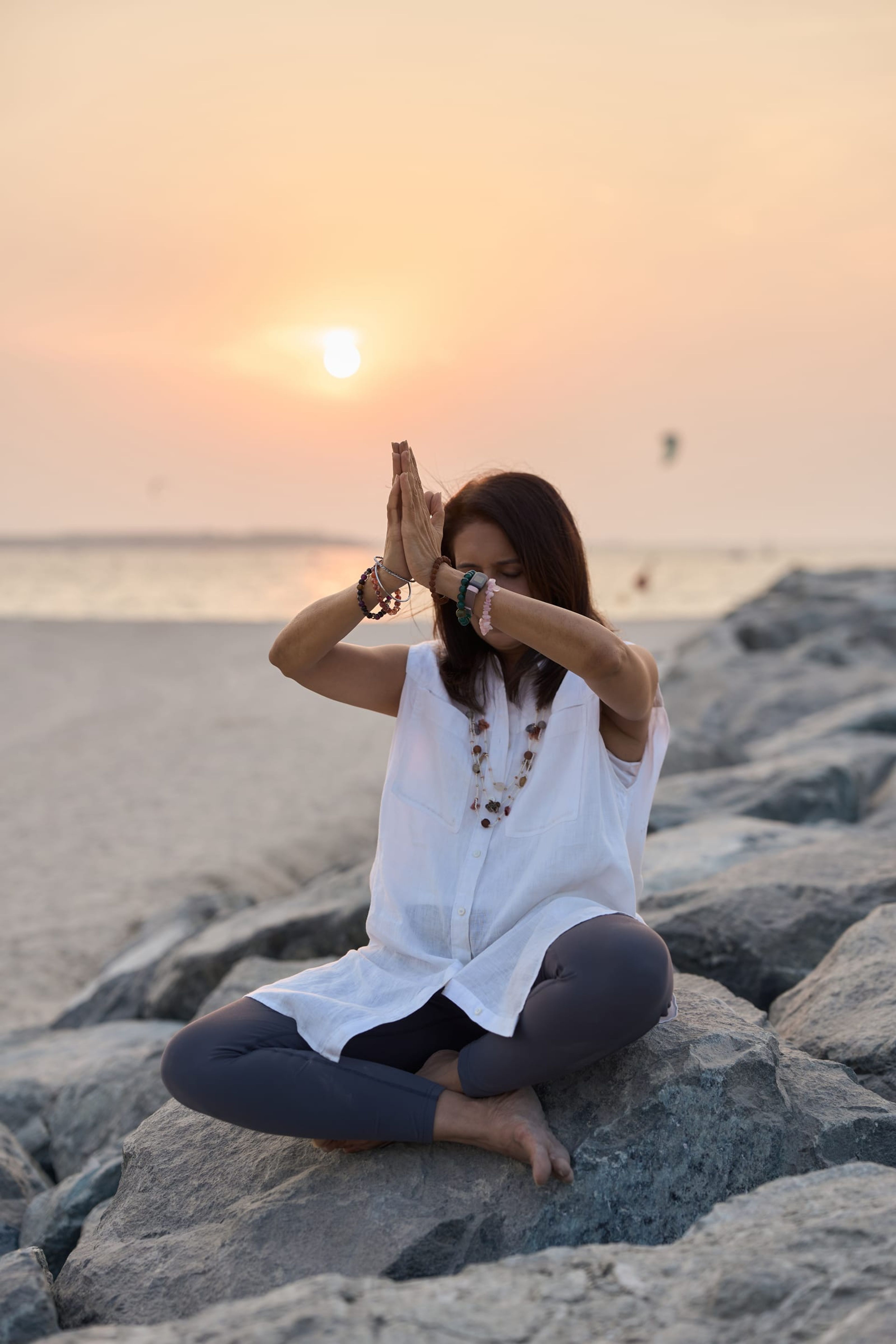 Manisha meditating on the rocks by the sea at sunset, hands joined above her head