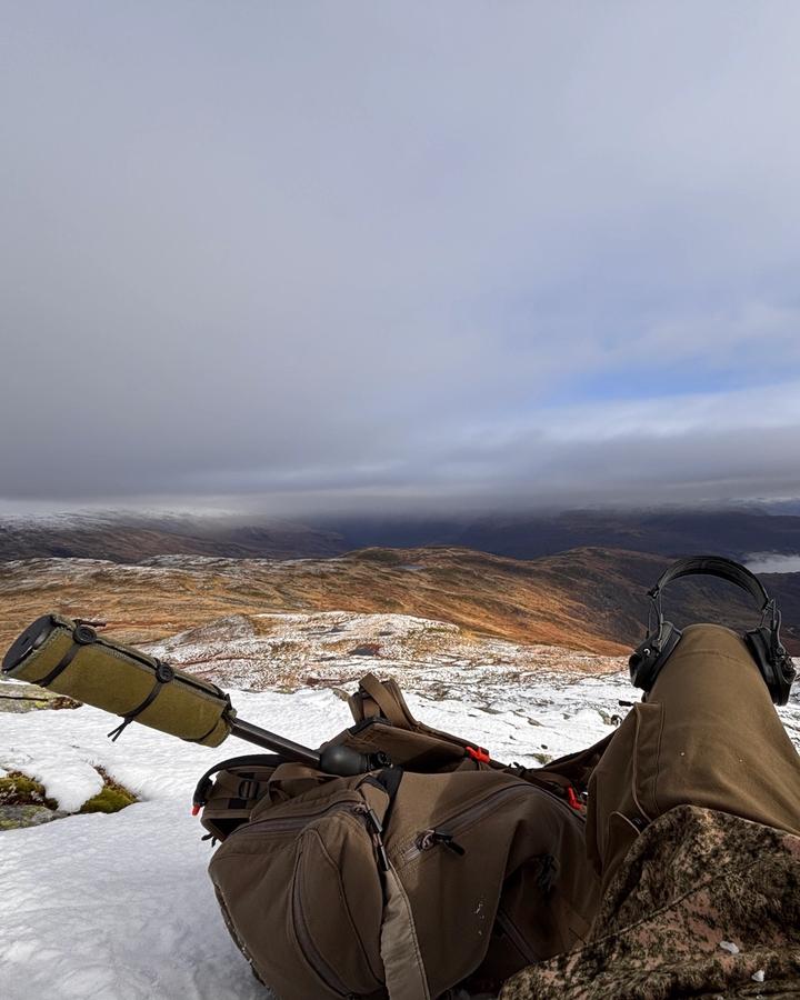 Utsikt fra en snødekt topp, med rifle, hørselvern og en oransje lue i forgrunnen.