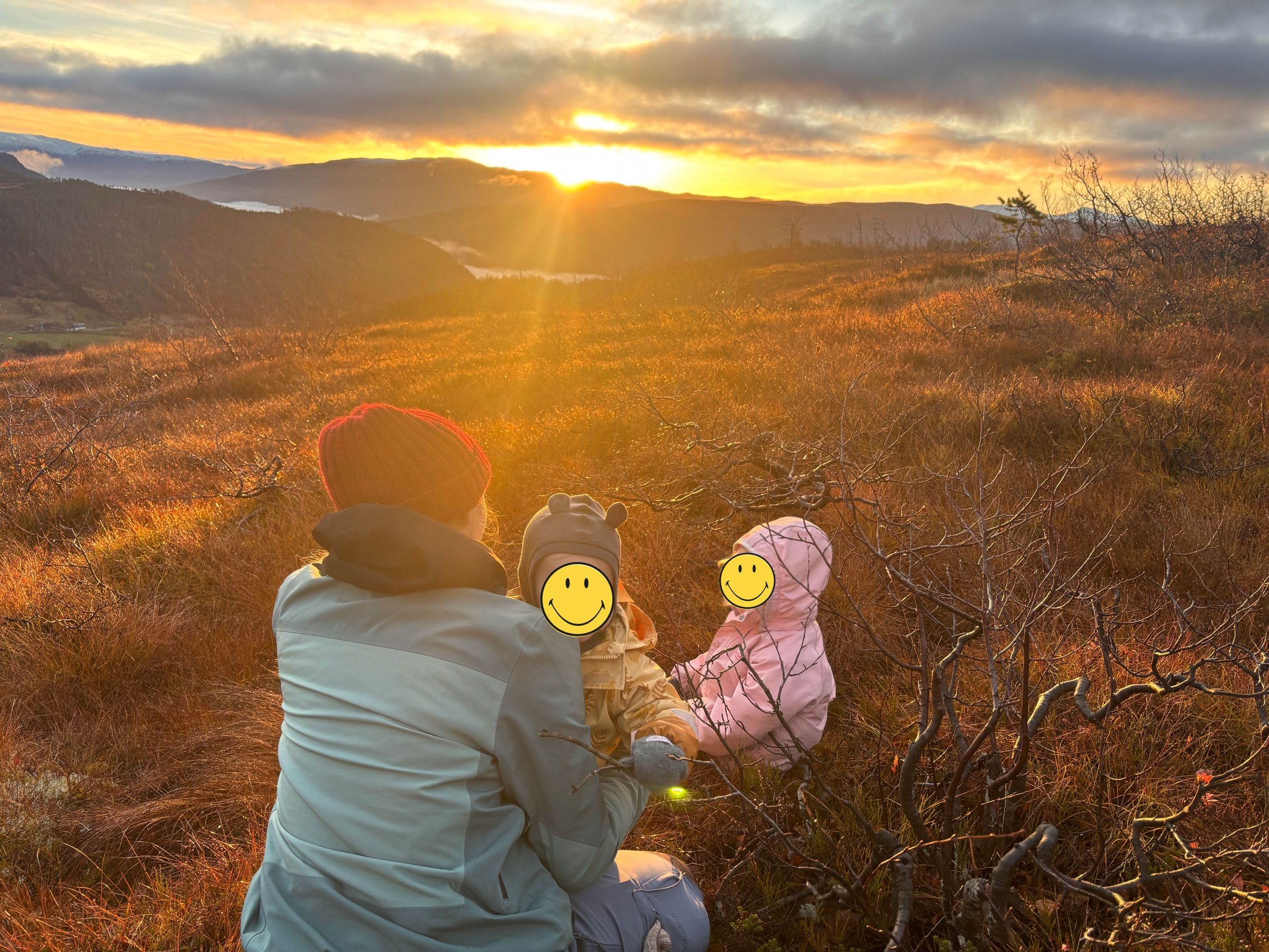 En voksen og to barn sitter i lyngen og ser på soloppgangen over åsene.