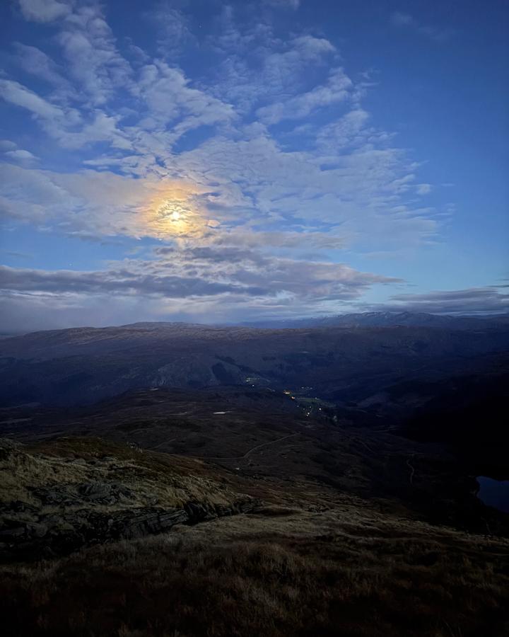 Månen bak skyer i blåtimen, med lys fra bygda nede i dalen.
