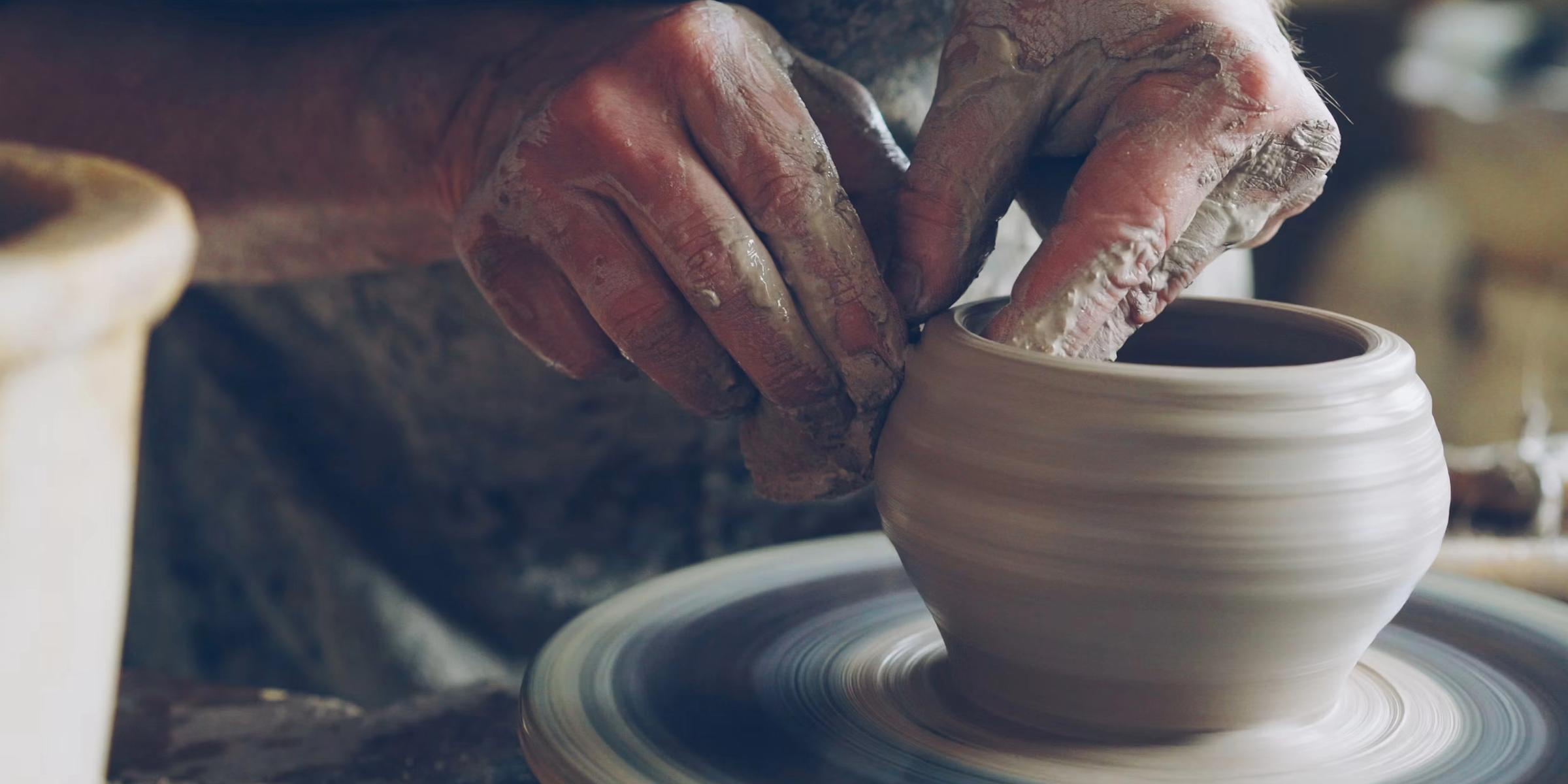 A potter's hands shaping wet clay on a spinning throwing wheel, centered and rising into a narrow vase.