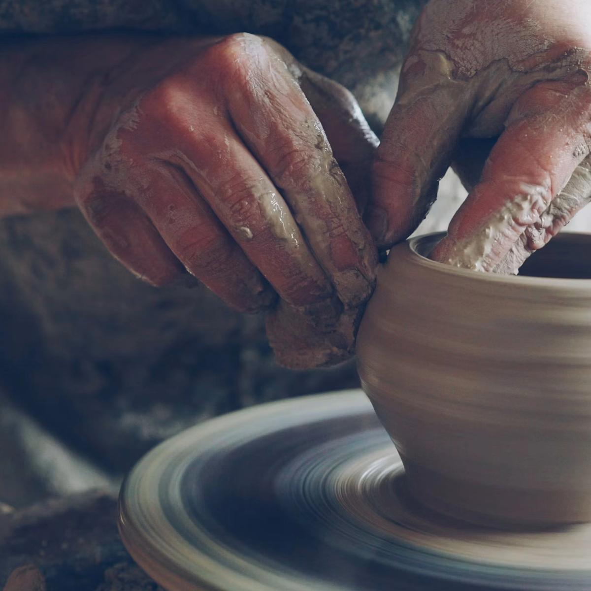 A potter's hands shaping wet clay on a spinning throwing wheel, centered and rising into a narrow vase.