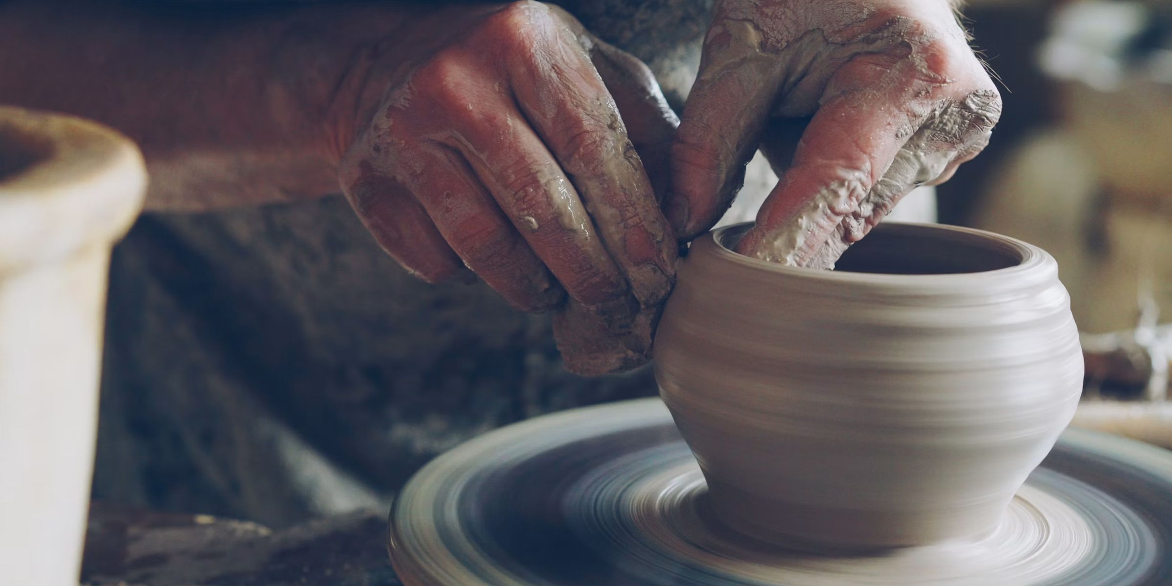 A potter's hands shaping wet clay on a spinning throwing wheel, centered and rising into a narrow vase.