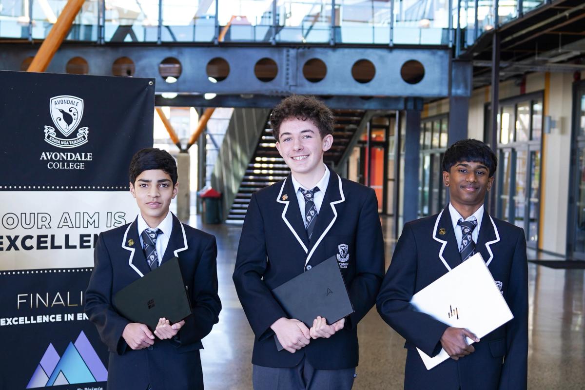 Three Avondale College students in school uniform holding laptops, in front of an Avondale College banner