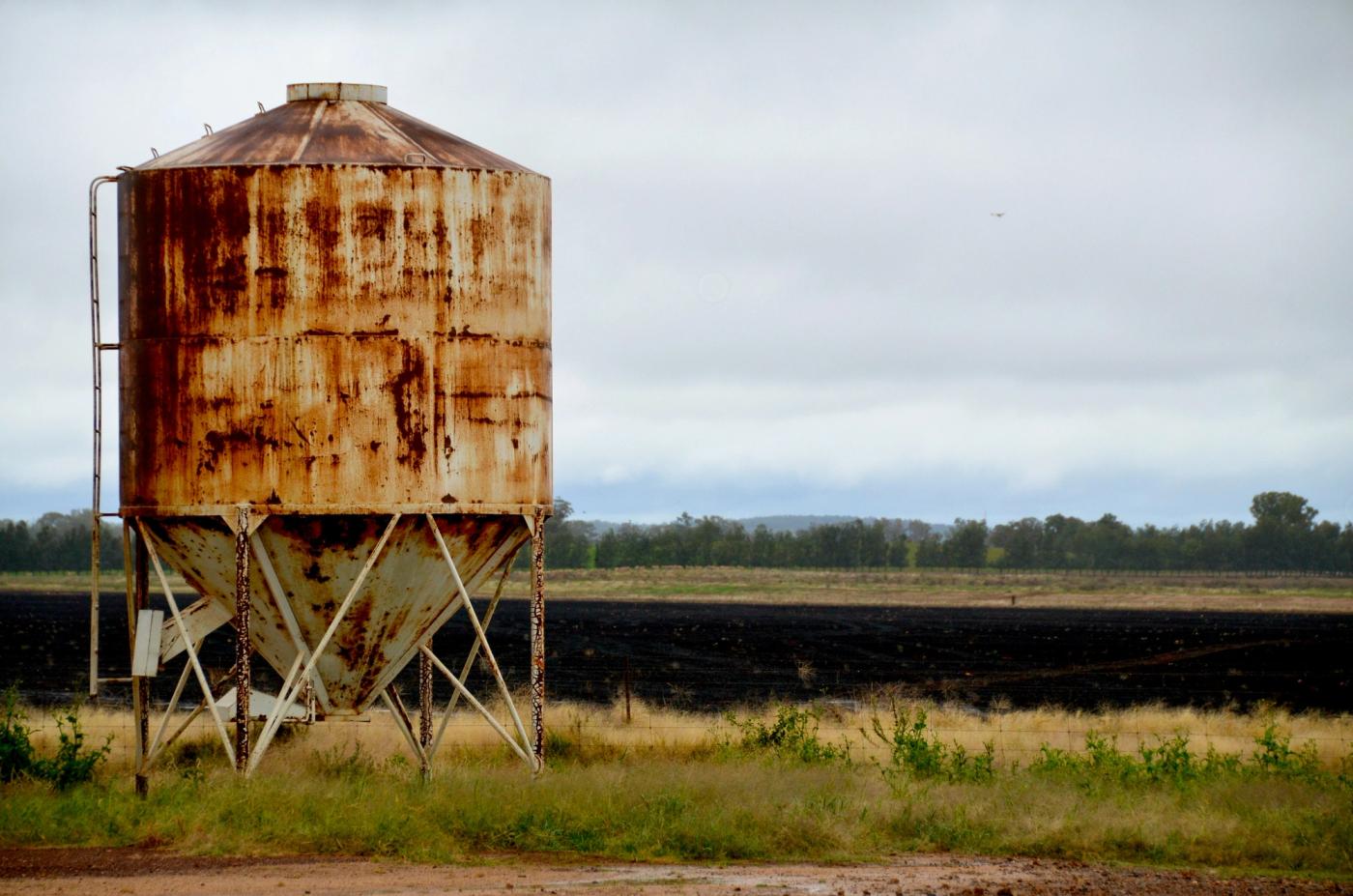 A weathered, rusting silo on its stand beside a farm paddock