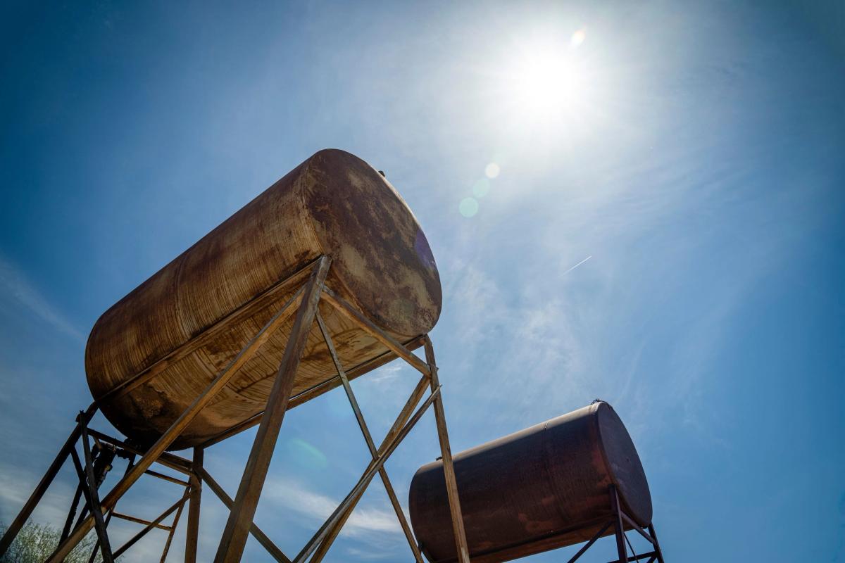 Two weathered overhead fuel tanks on steel stands, seen from below against a blue sky