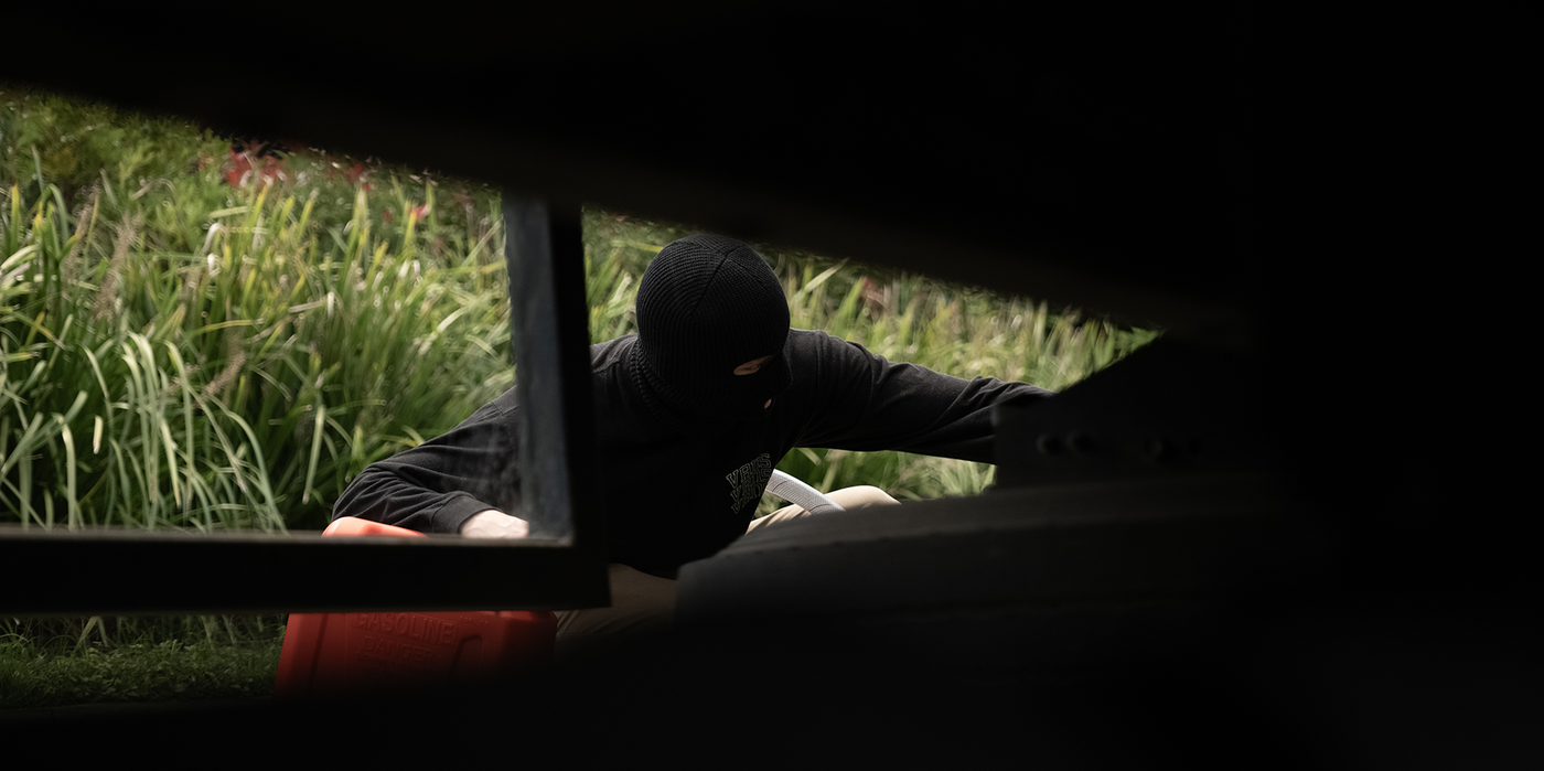 A masked figure syphoning fuel from a parked vehicle at night