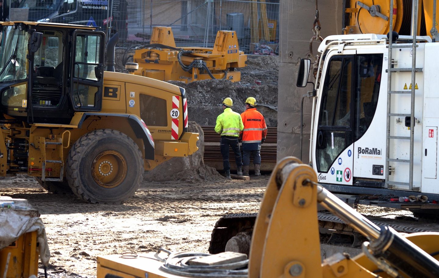 Two workers in hi-vis between a wheel loader and a piling rig on a construction site