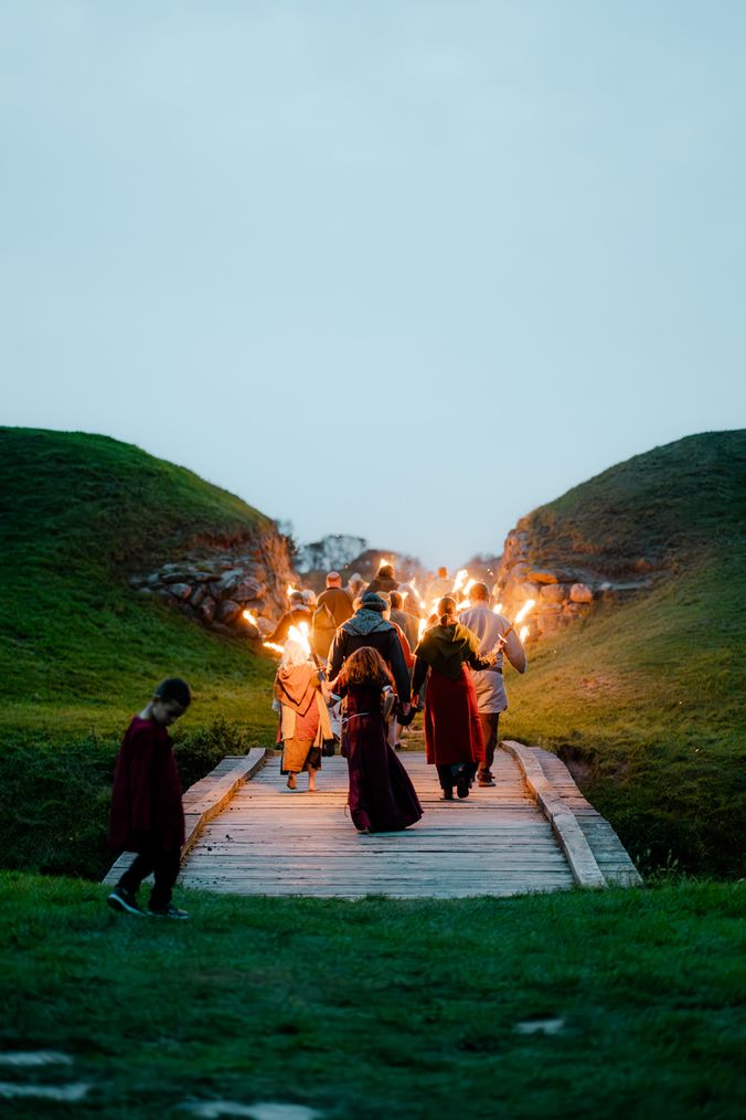 Group of people in Viking clothing walking on wooden bridge toward the ring fortress at dusk with torches in their hands.