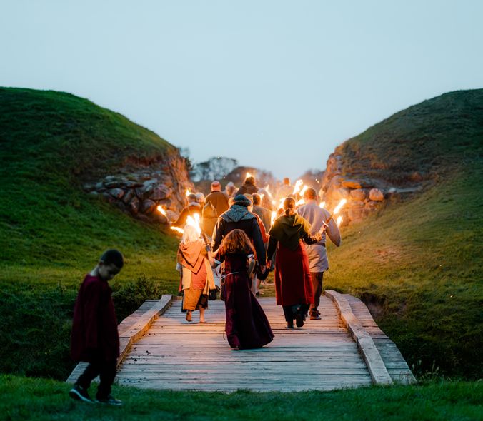 Gruppe mennesker i vikingetøj går på træbro ind mod ringborgen ved aftenstid med fakler i hænderne.