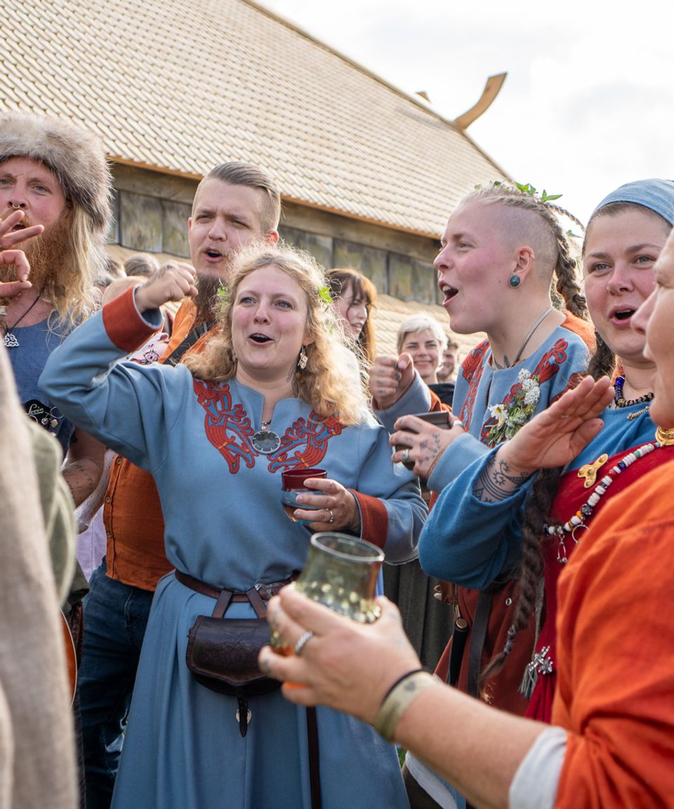 Festklædte deltagere i farverige vikingedragter fejrer et vikingebryllup foran langhuset på Trelleborg.