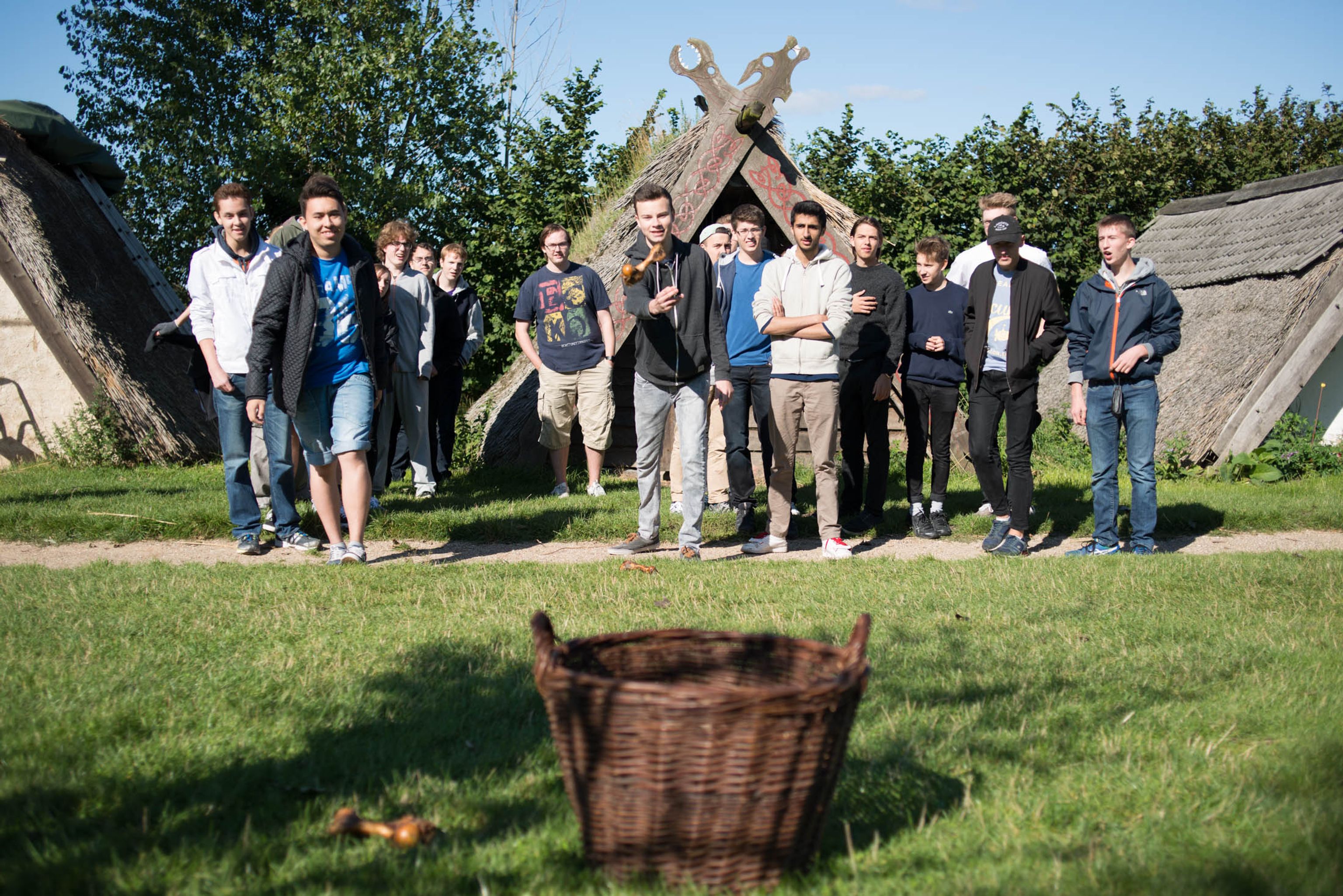 Group of young men preparing a throwing exercise with a woven basket as the target in front of Viking houses at Trelleborg.