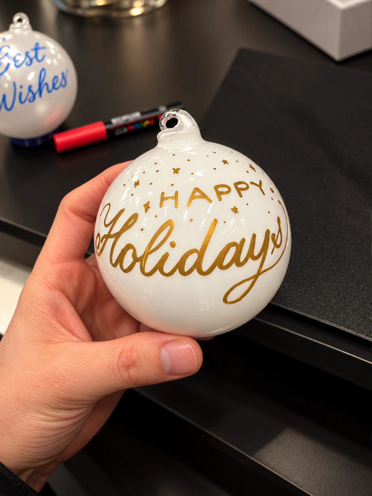 Hand holding a white ornament with gold Happy Holidays lettering