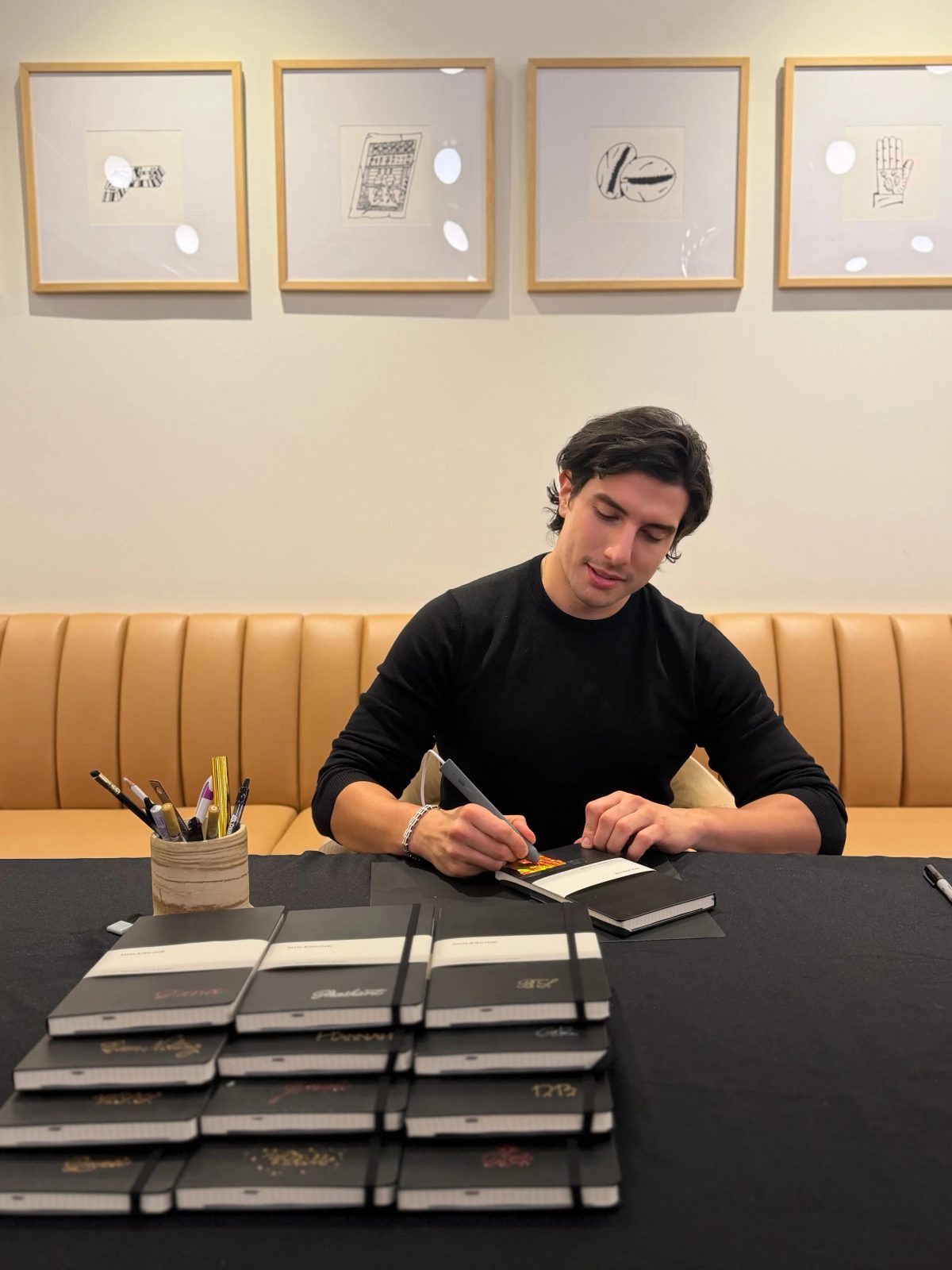 Artist lettering a small notebook at a table of black notebooks