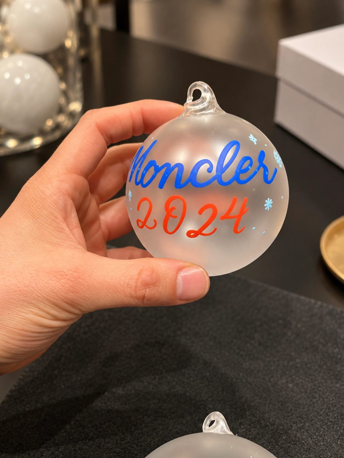 Hand holding a white ornament with blue and red Moncler 2024 lettering
