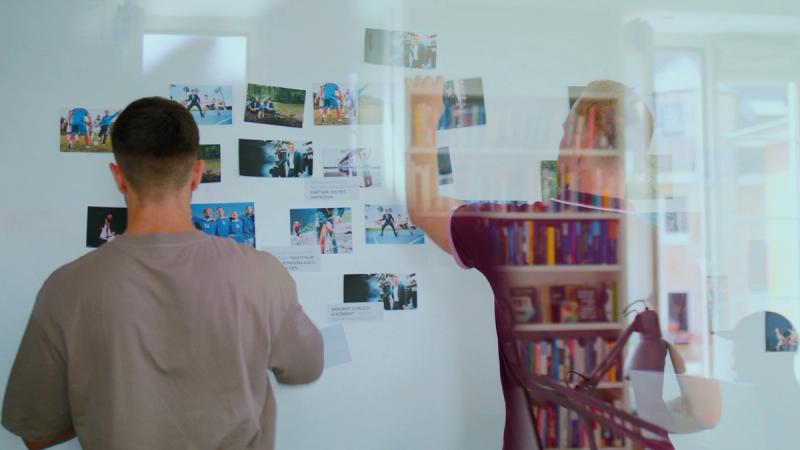 Zwei Personen ordnen Fotos und Notizen für ein Moodboard an einer Glaswand im Büro.