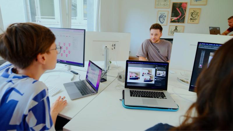 Mehrere Personen arbeiten gemeinsam in einem hellen Büro an digitalen Designentwürfen auf Laptops und großen Bildschirmen.