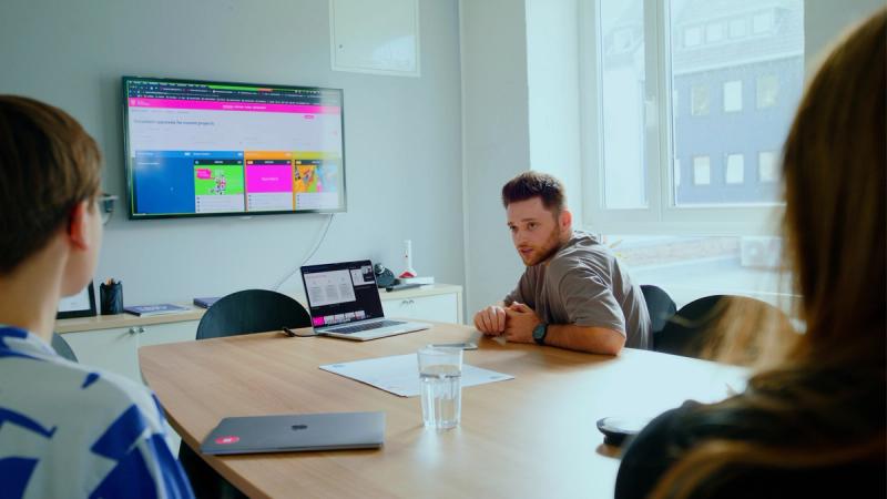 Drei Personen sitzen in einem Meetingraum an einem Tisch und betrachten gemeinsam ein Brandportal auf einem großen Bildschirm.