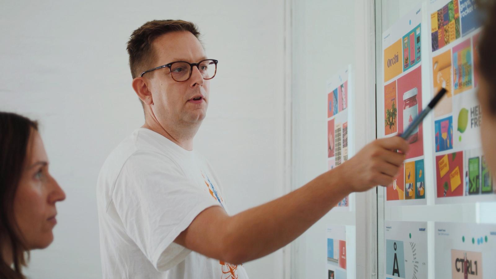 Ein Mann präsentiert mit einem Stift verschiedene farbige Designentwürfe an einer Wand vor zwei weiteren Personen.
