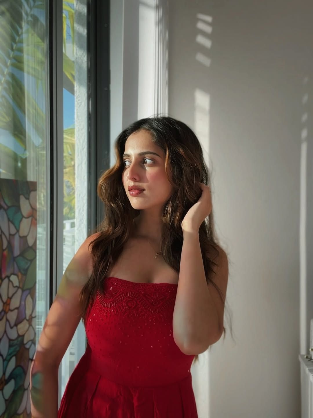 Creator Nivedita Gowda wearing a stylish red outfit, standing by a sunlit window with soft natural lighting