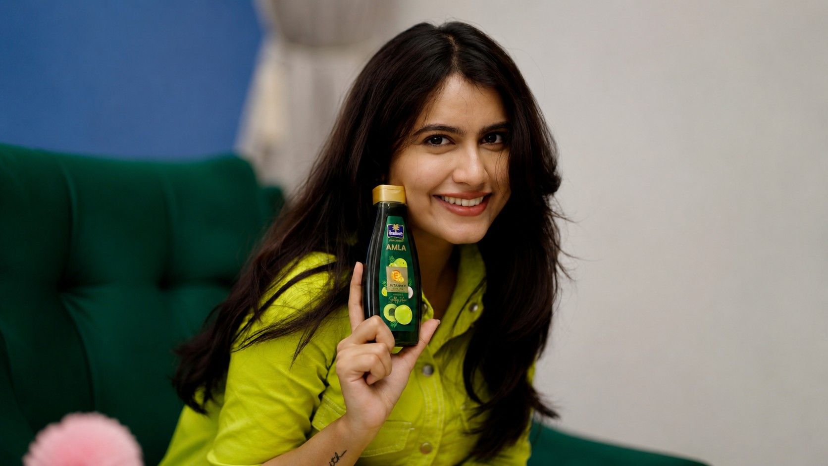 Creator Bunny Vox smiling while holding a Parachute Advansed Amla hair oil bottle, wearing a bright green outfit in a cozy indoor setting