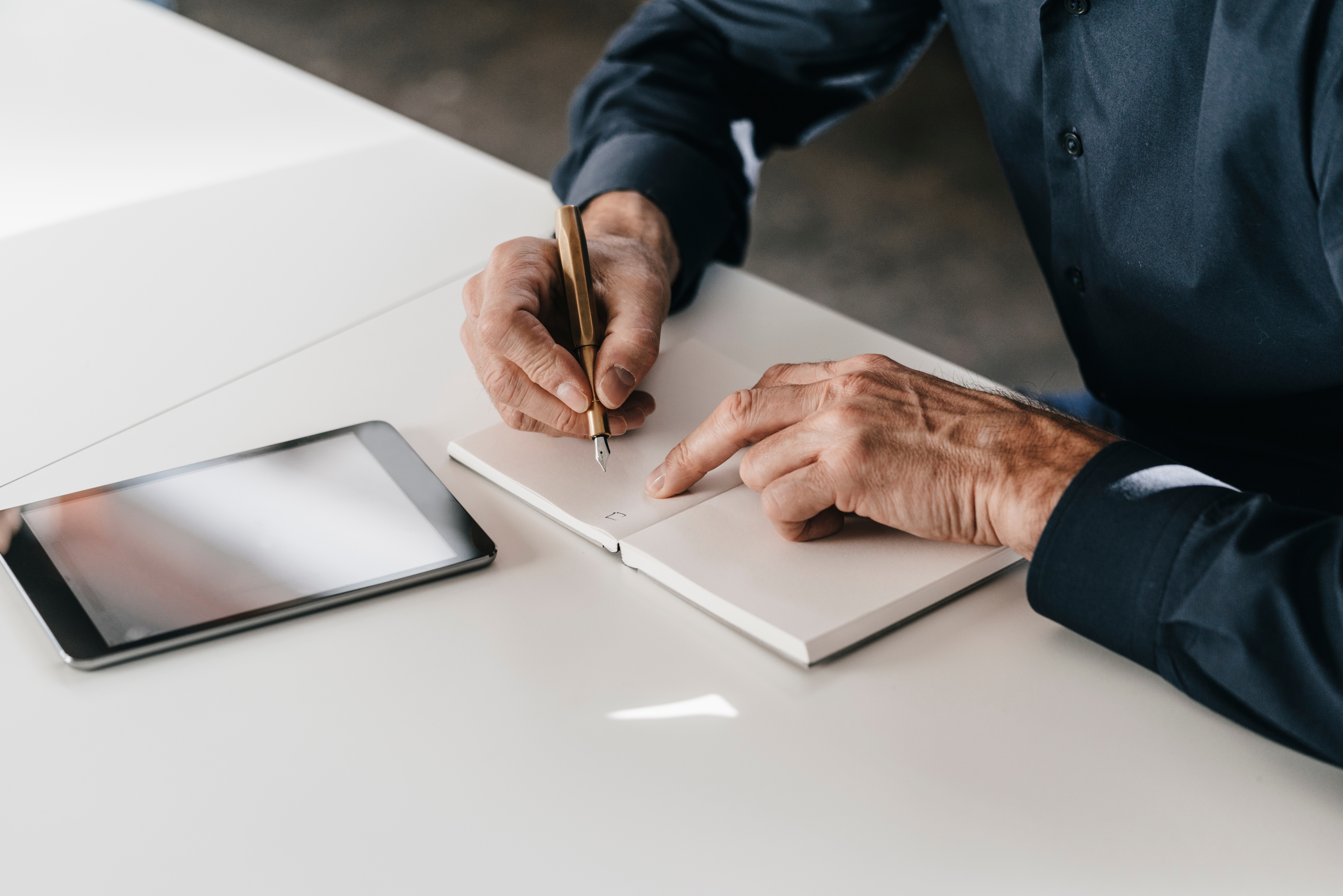 A person writes in a notebook beside a tablet on a desk.