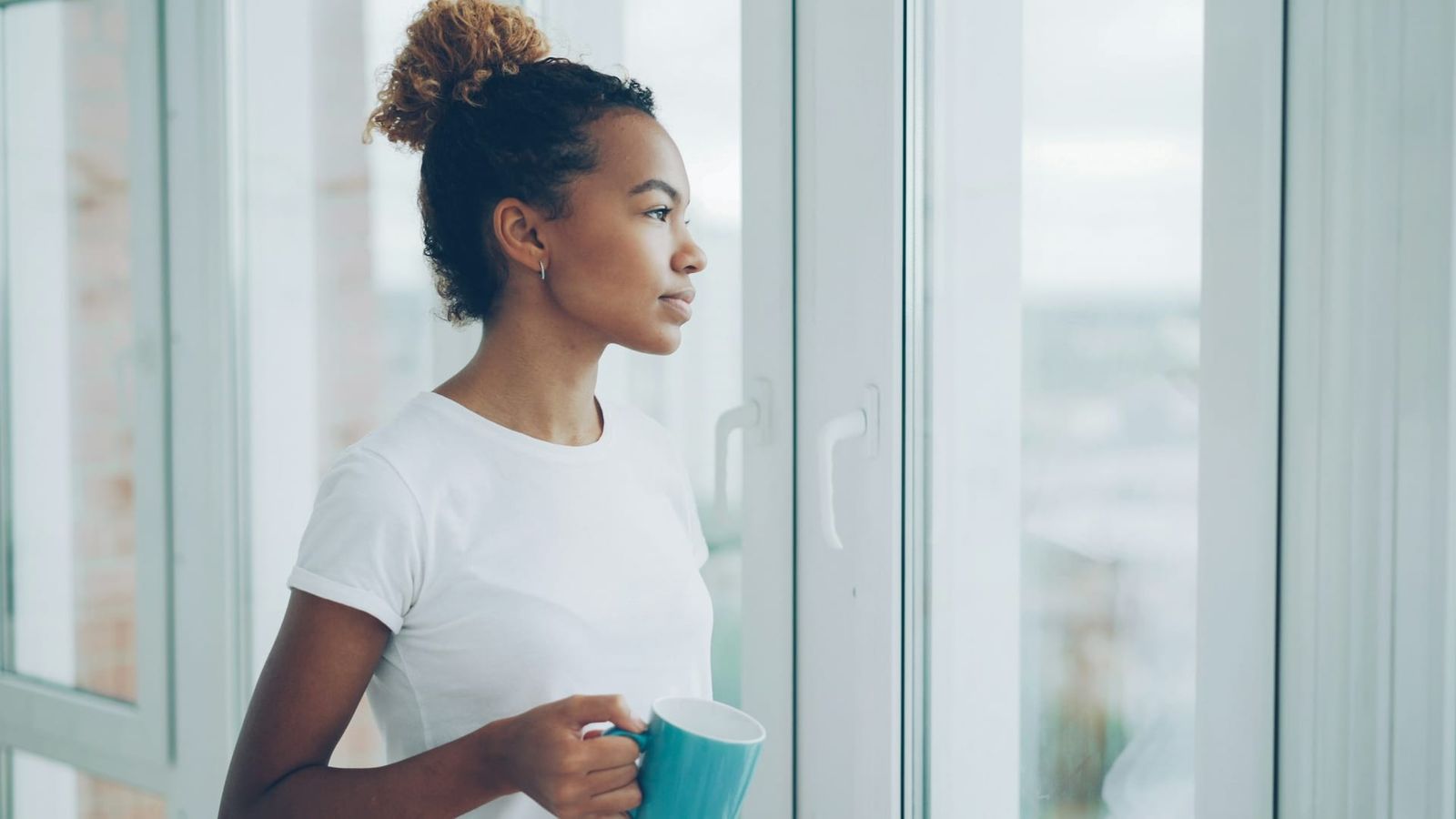 A person standing quietly at a window with a warm mug, looking out