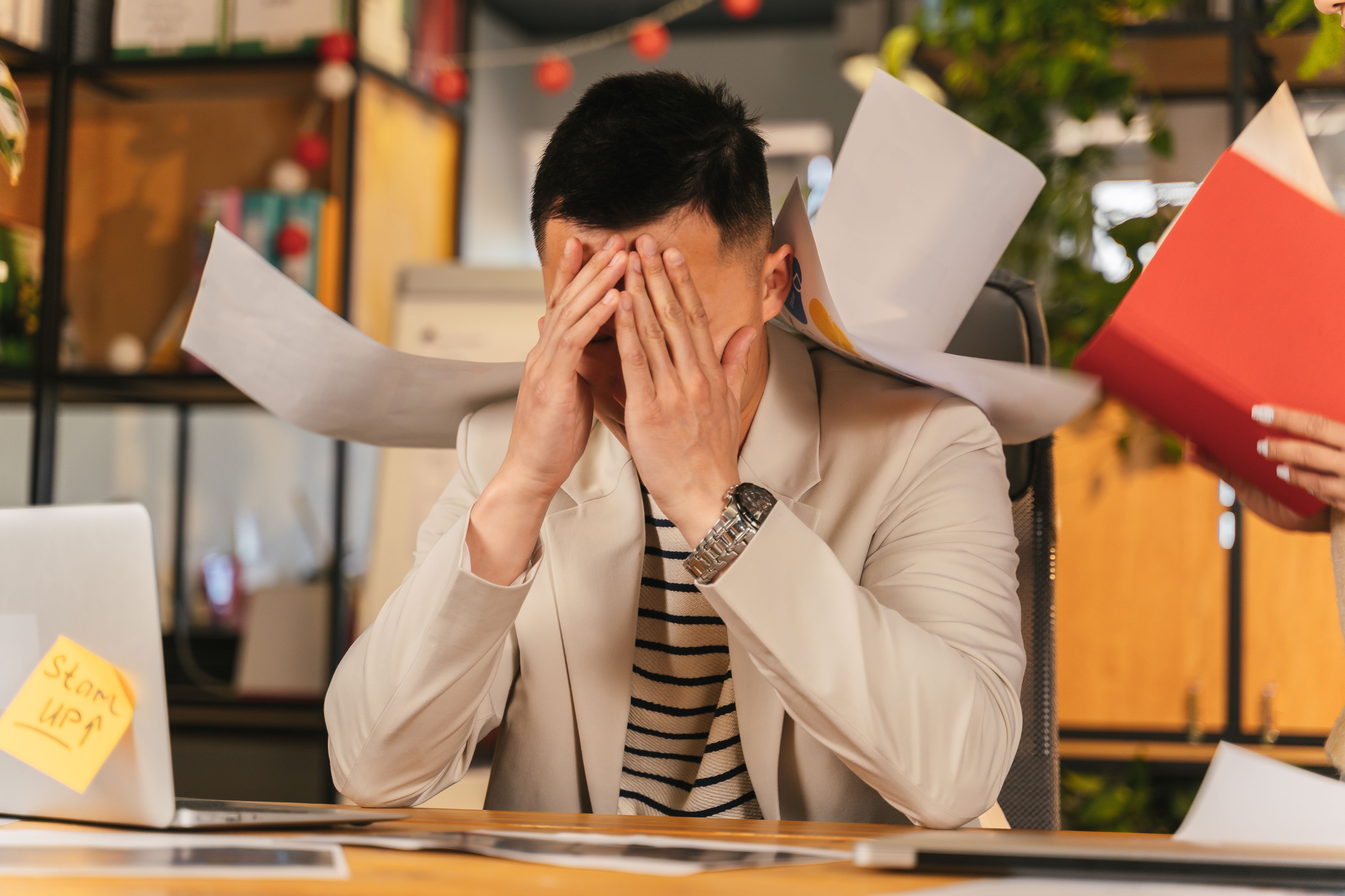 A stressed office worker in a beige blazer covers his face with both hands at a desk while papers fly around him and a colleague holds out a red folder.