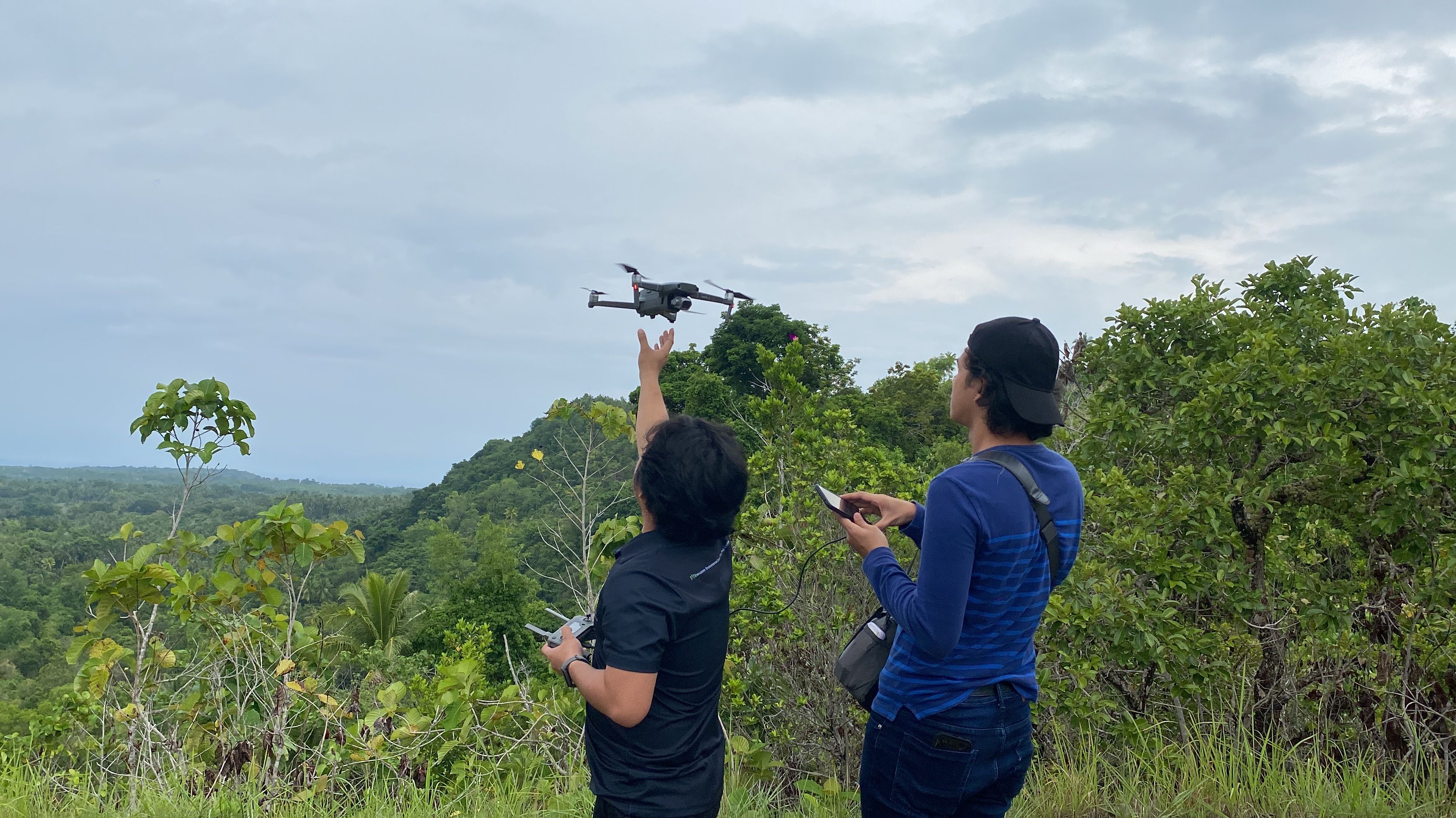 Two Envi-Comm field team members on a forested hilltop launching a drone, one releasing it by hand while the other flies it from a phone.