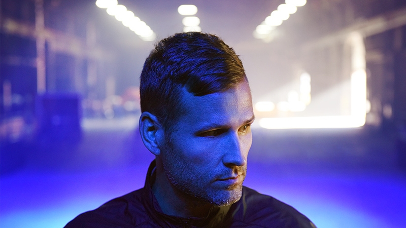 Kaskade with a blue light on his face and a warehouse in the background