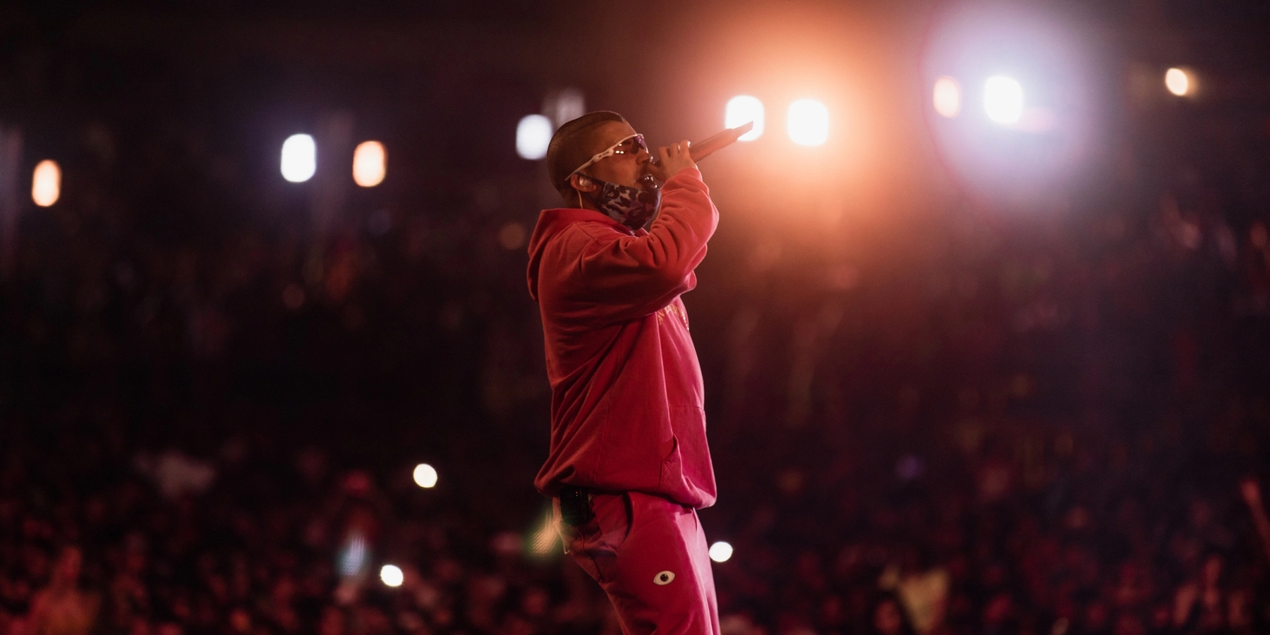 Bad Bunny on the Baja Beach Fest stage holding a microphone up to his face. He is wearing a pink hoodie, and pink sweat pants.