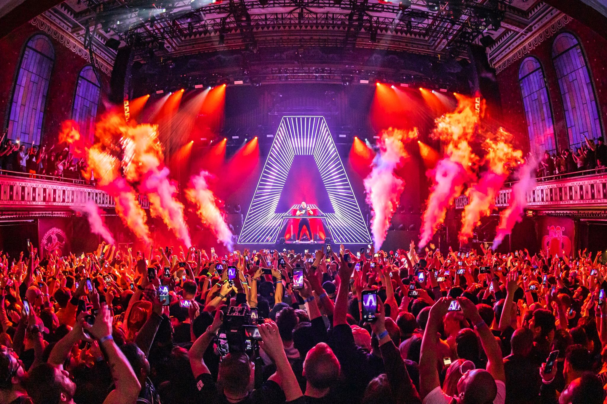 Crowd point of view shot of Armin Van Buuren performing in front of a packed crowd as CO2 bursts from either side of his "A" production DJ booth.