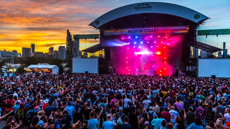 Large crowd gathered in front of the North Coast stage with purple, red and yellow lights illuminating the fog on the stage. The sun is setting on the Chicago skyline, as the sky is partly yellow and orange and blue.