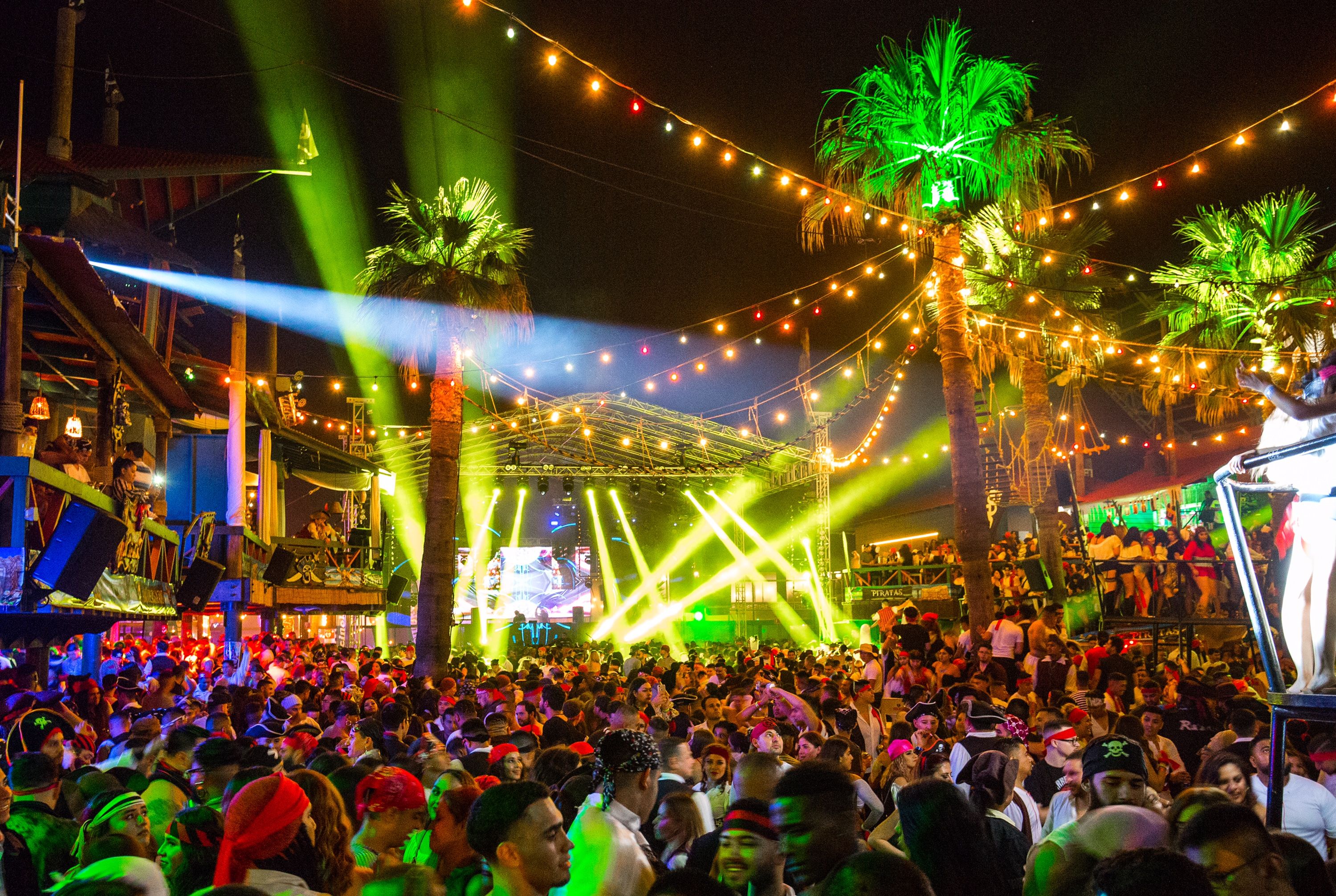 A wide angle shot of Papas & Beer located at Rosarito, MX with large palm trees, a large crowd, and a big main stage with yellow production lights.