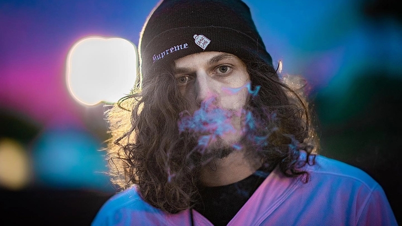 Jesse Kardon aka Subtronics wearing a black beanie, a purple shirt, and a black under shirt blowing smoke towards the camera. The photo has a vibrant blue purple tint.