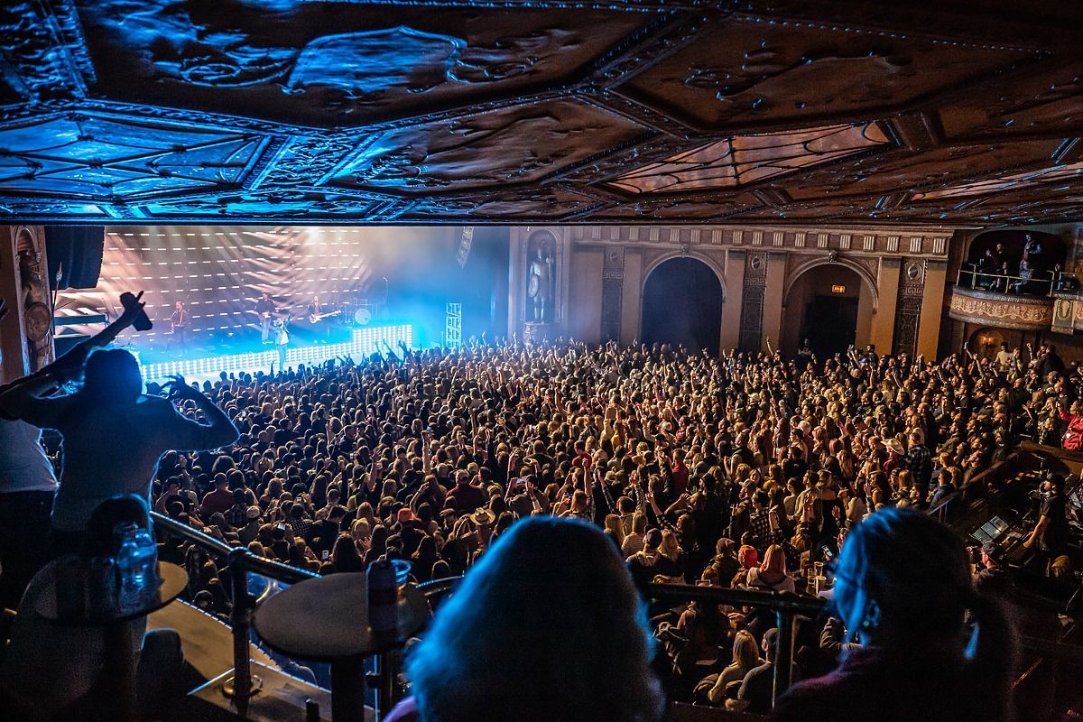 A photo shot from the back left area of the venue on the 2nd floor. The production lights are blue, and a large crowd is gathered in front of the main stage. 
