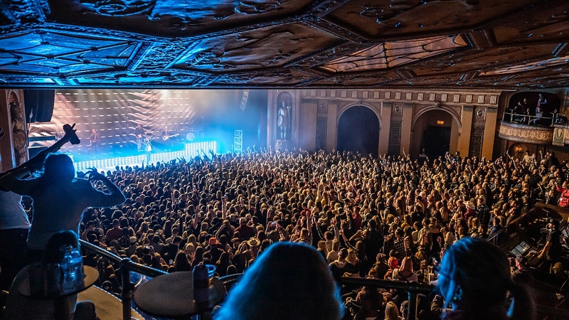 A photo shot from the back left area of the venue on the 2nd floor. The production lights are blue, and a large crowd is gathered in front of the main stage.