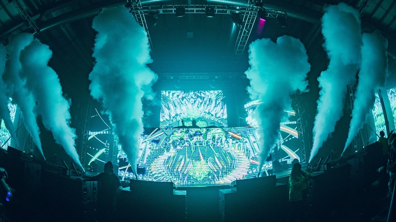 Front of a stage illuminated with blue light and CO2 shooting off the front of the stage with a dj booth behind it