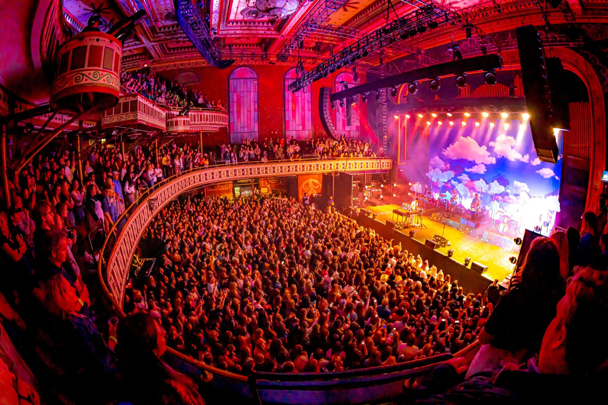 A view from the right side balcony facing the Tabernacle stage showcasing a large packed crowd on the first, second, and third floors. The production lights give an orange glow to the crowd. 