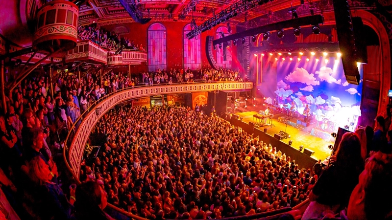 A view from the right side balcony facing the Tabernacle stage showcasing a large packed crowd on the first, second, and third floors. The production lights give an orange glow to the crowd.