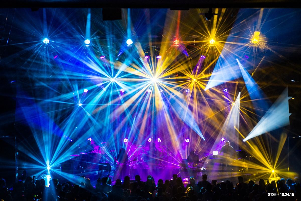 Left to right: Blue lights, yellow lights, white lights highlight the main stage of the Coca-Cola Roxy venue as a crowd is gathered in front. 