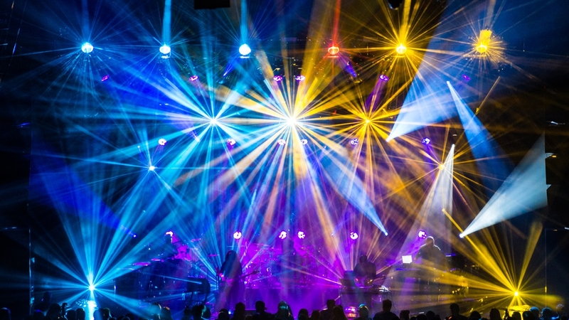 Left to right: Blue lights, yellow lights, white lights highlight the main stage of the Coca-Cola Roxy venue as a crowd is gathered in front.