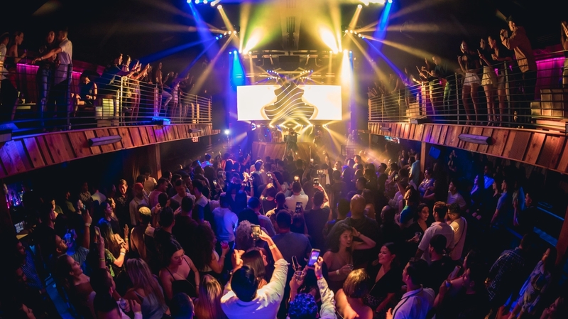 Wide BOH shot of a full crowd at Ravine as an artist performs. The crowd is also on either side on the balconies as the production lights shine yellow and blue on the crowd