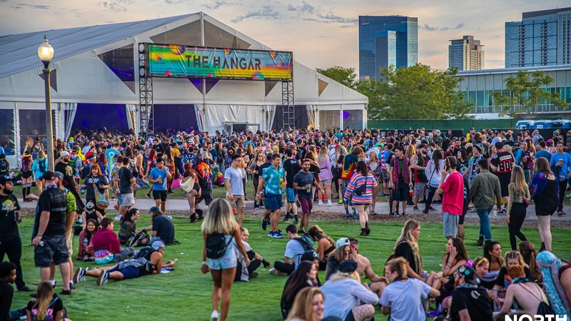 A lot of people hanging out and or walking by a large white tent labeled "The Hangar" at North Coast Music Festival. A few skyscraper buildings line the right side of the photo.