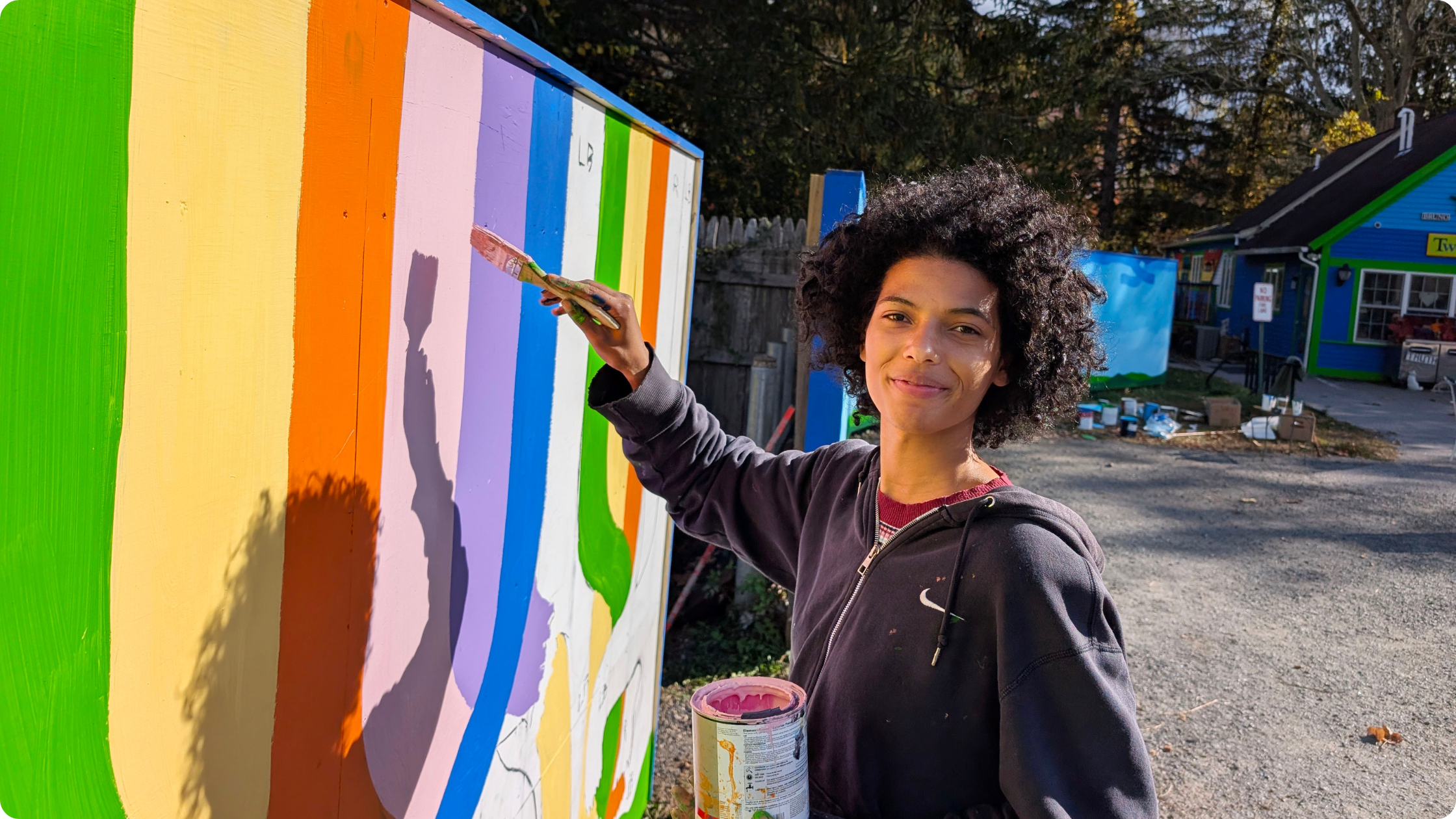 A person with medium skin tone and an afro wearing a dark Nike zip-up hoodie paints a mural featuring vibrant colors.