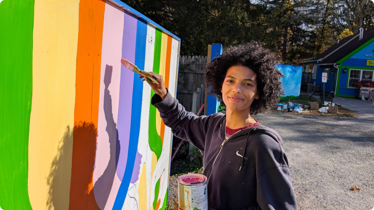 A person with medium skin tone and an afro wearing a dark Nike zip-up hoodie paints a mural featuring vibrant colors.