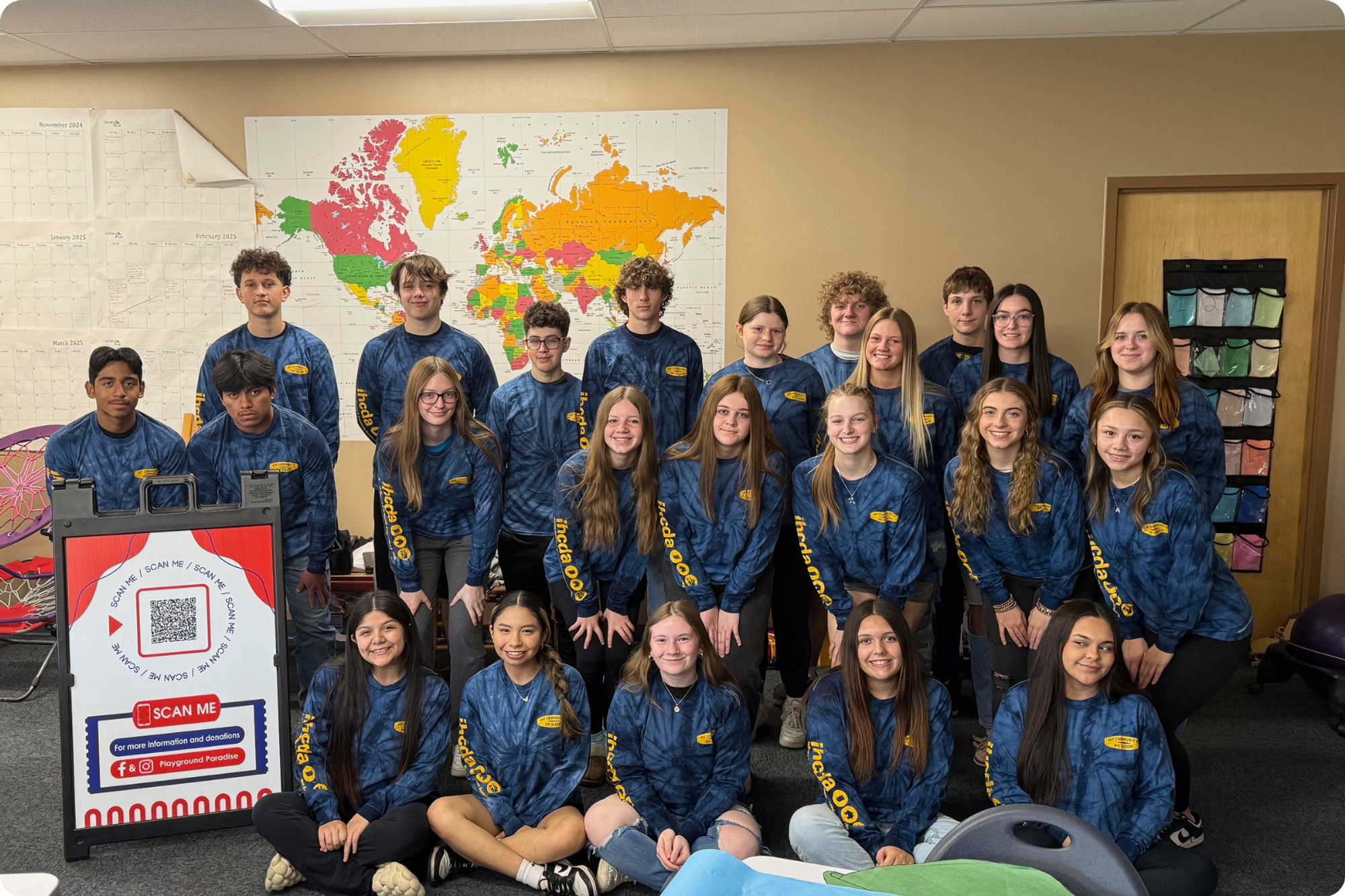 23 teenagers pose for a group photo in a classroom, wearing matching blue IHCDA shirts