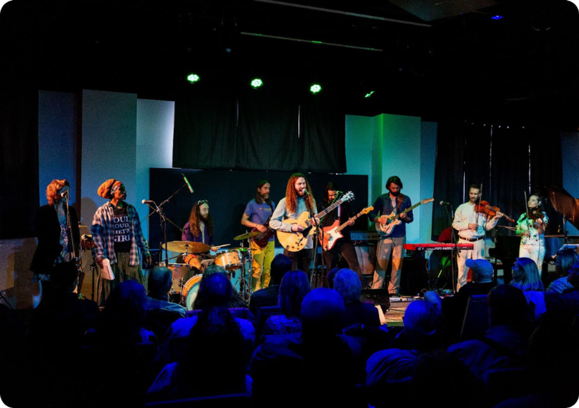 9 musicians on an indoor stage perform for a seated crowd