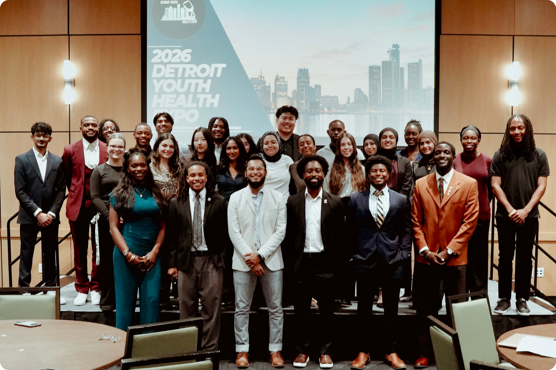 Three rows of university students pose in front of a projection screen that reads "2026 Detroit Youth Health Expo"