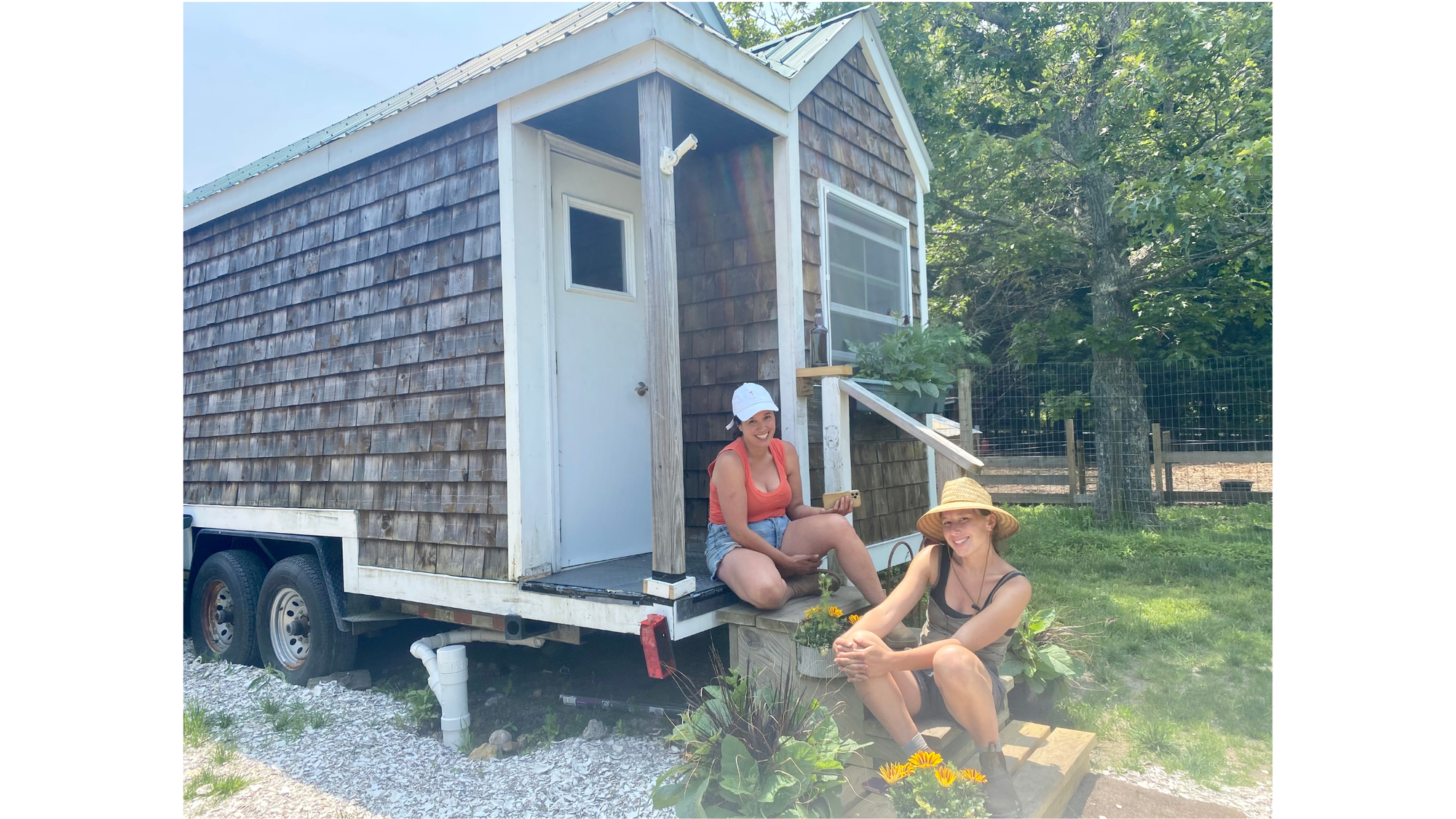 Two women smile for  the camera while sitting on the steps of a small mobile shed. 
